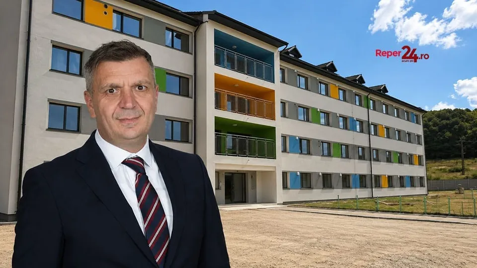 Un bărbat într-un costum închis și cravată în dungi stă în prim-plan în fața unui bloc de apartamente cu ferestre colorate sub un cer senin.