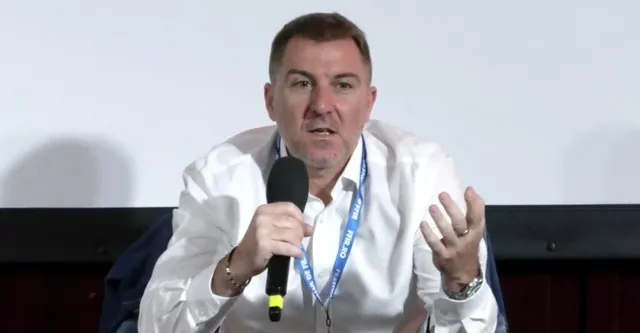 Un bărbat cu cămașă albă și lanyard albastru vorbește într-un microfon ținut cu mâna dreaptă, având mâna stângă ridicată în gest.