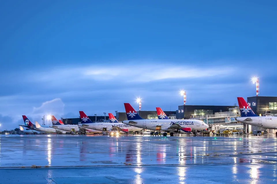 Mai multe avioane de pasageri cu marcaje roșii și albastre sunt parcate pe o pistă de aeroport umedă sub un cer albastru.