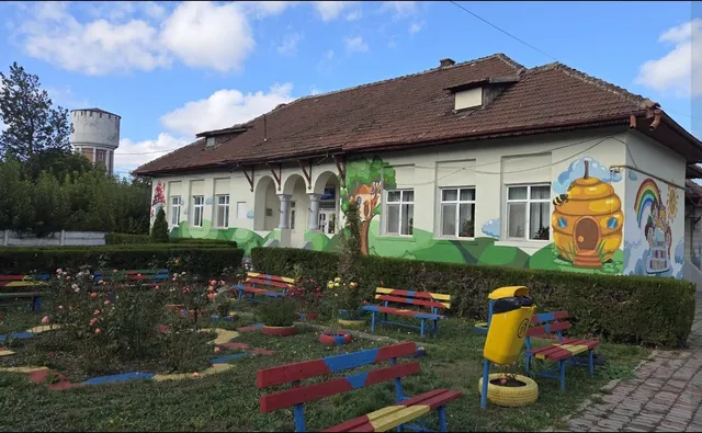 O clădire cu fațadă decorată cu desene colorate este situată lângă o grădină cu bănci de culori diferite și un loc de joacă galben.