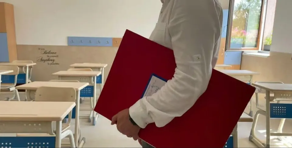 O persoană într-o cămașă albă poartă două scaune de culoare roșie într-o încăpere cu bănci de școală albe și albastre.