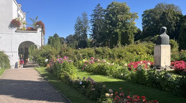 Un drum pavat trece pe lângă straturi de flori colorate și un bust pe un soclu înalt într-o grădină cu copaci verzi.
