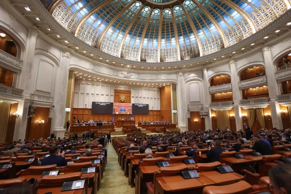 O sală de conferințe circulară cu scaune de lemn dispuse în rânduri sub un dom înalt cu ferestre decorate și panouri digitale pe perete.