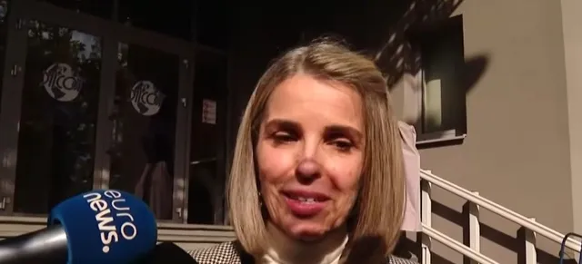 O femeie cu părul blond și bluză albă vorbește în fața unui microfon albastru cu inscripția euro news.