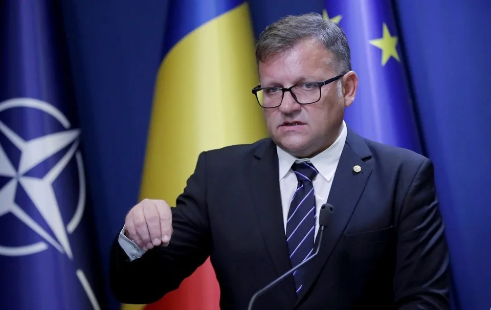 Un bărbat cu ochelari, purtând costum închis la culoare și cravată striată, stă în fața unor steaguri pe un fundal albastru.