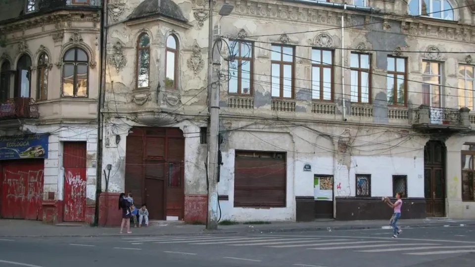 O clădire veche cu ferestre înalte și fațadă deteriorată se află lângă un drum asfaltat cu marcaje de trecere de pietoni.