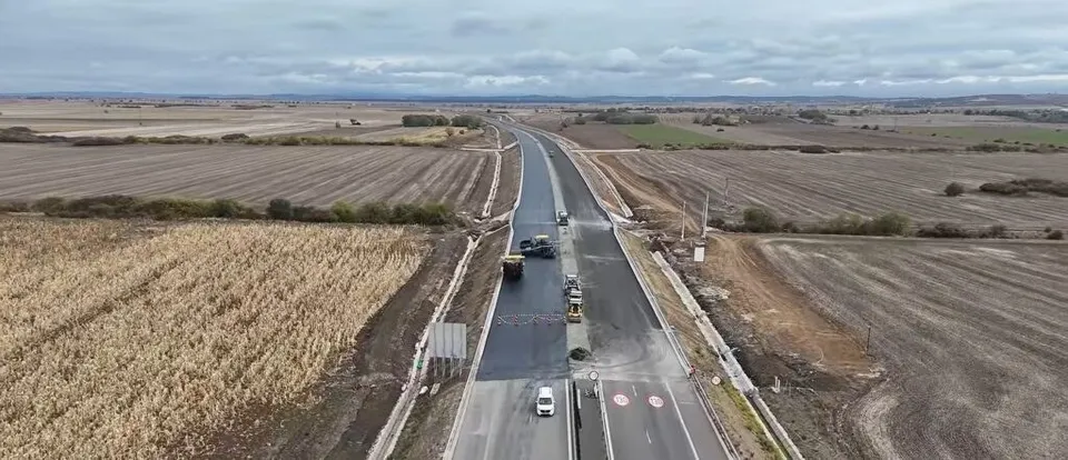 O vedere aeriană prezintă o porțiune de autostradă cu benzi de asfalt negru și gri, înconjurată de câmpuri agricole deschise sub un cer înnorat.