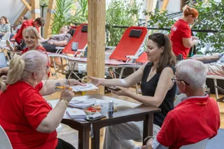 O femeie în vestimentație neagră stă la o masă lângă o persoană în tricou roșu, în timp ce o altă femeie în tricou roșu stă pe un scaun.