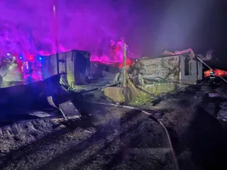 Structuri de zidărie sunt afectate de flăcări și fum sub o lumină purpurie intensă pe un teren de noapte.