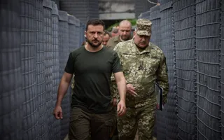 Un bărbat cu tricou verde închis și pantaloni militari merge printr-un coridor format din bariere de protecție gri, însoțit de mai mulți bărbați îmbrăcați în uniforme militare.