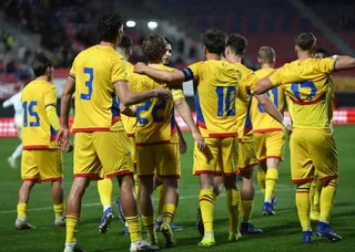 Un grup de fotbaliști în echipament galben cu detalii albastre și roșii stau întruniți pe un teren de fotbal sub luminile unui stadion.