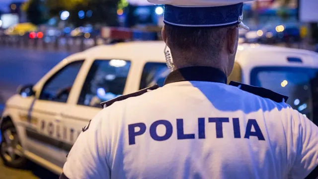Un polițist cu inscripția POLITIA pe uniformă stă cu spatele, privind un autovehicul alb parcat pe un drum urban noaptea.