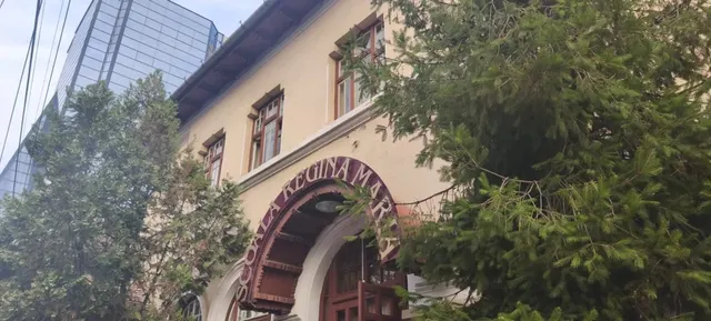 O clădire cu ferestre maronii și un arc de intrare pe care scrie text, parțial acoperită de copaci verzi.