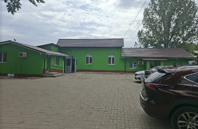 O clădire verde cu acoperiș gri este situată lângă o curte pavată unde sunt parcate câteva mașini sub un cer înnorat.