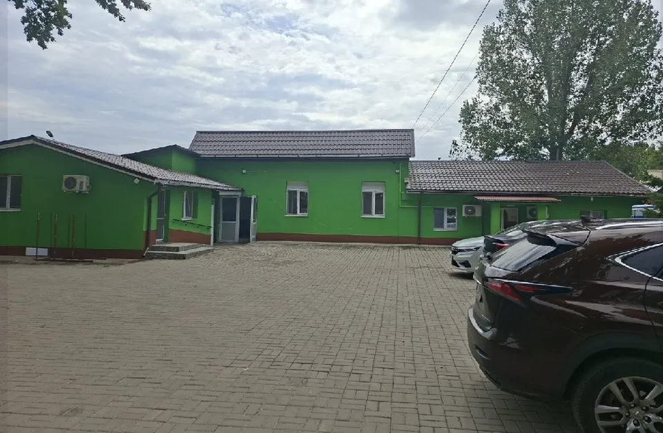 O clădire verde cu acoperiș gri este situată lângă o curte pavată unde sunt parcate câteva mașini sub un cer înnorat.