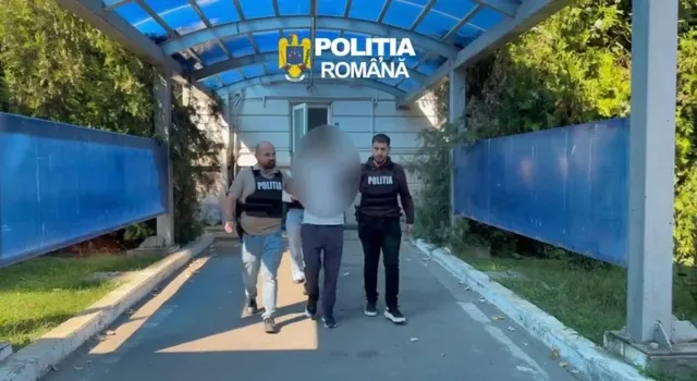 Trei persoane merg pe un drum asfaltat între garduri albastre, două dintre ele purtând veste negre cu inscripții albe.