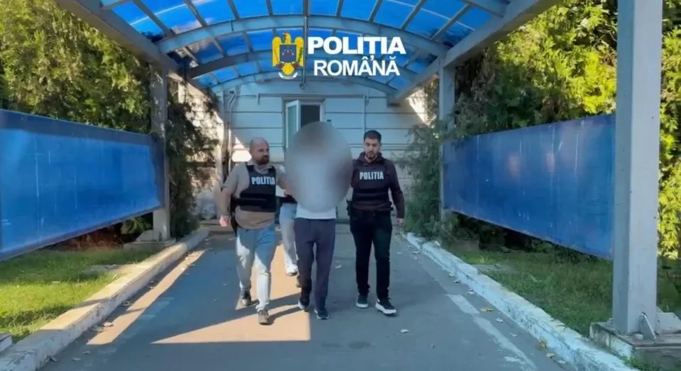Trei persoane merg pe un drum asfaltat între garduri albastre, două dintre ele purtând veste negre cu inscripții albe.