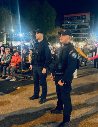 Doi bărbați în uniforme închise de poliție stau în picioare pe o stradă luminată noaptea, în timp ce mulțimea stă pe scaune în fundal.