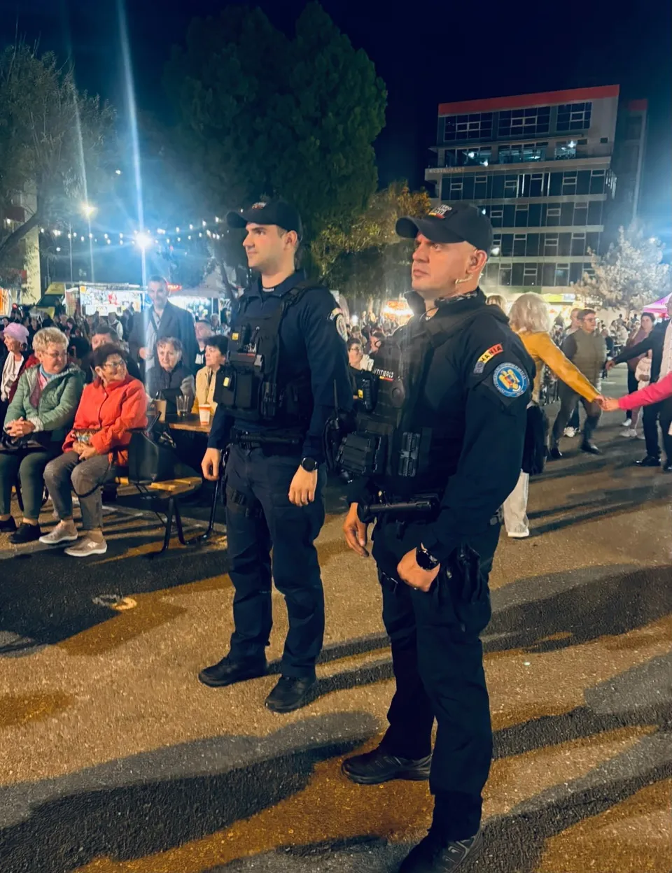 Doi bărbați în uniforme închise de poliție stau în picioare pe o stradă luminată noaptea, în timp ce mulțimea stă pe scaune în fundal.