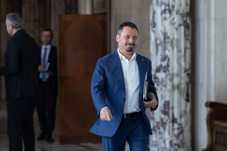 Un bărbat cu barbă și sacou albastru merge printr-un hol, purtând o cămașă albă și un dosar sub braț.