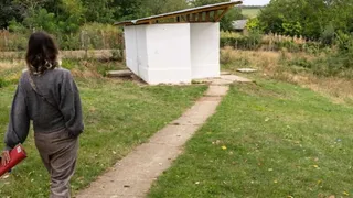 O femeie cu părul închis la culoare merge pe un drum îngust de beton spre o clădire mică, albă, cu acoperiș înclinat.