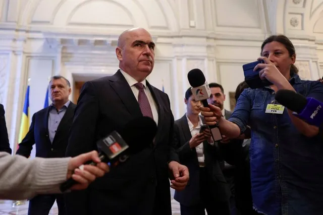 Un bărbat într-un costum închis și cravată purpurie este înconjurat de microfoane și persoane în interiorul unei clădiri cu arhitectură clasică.