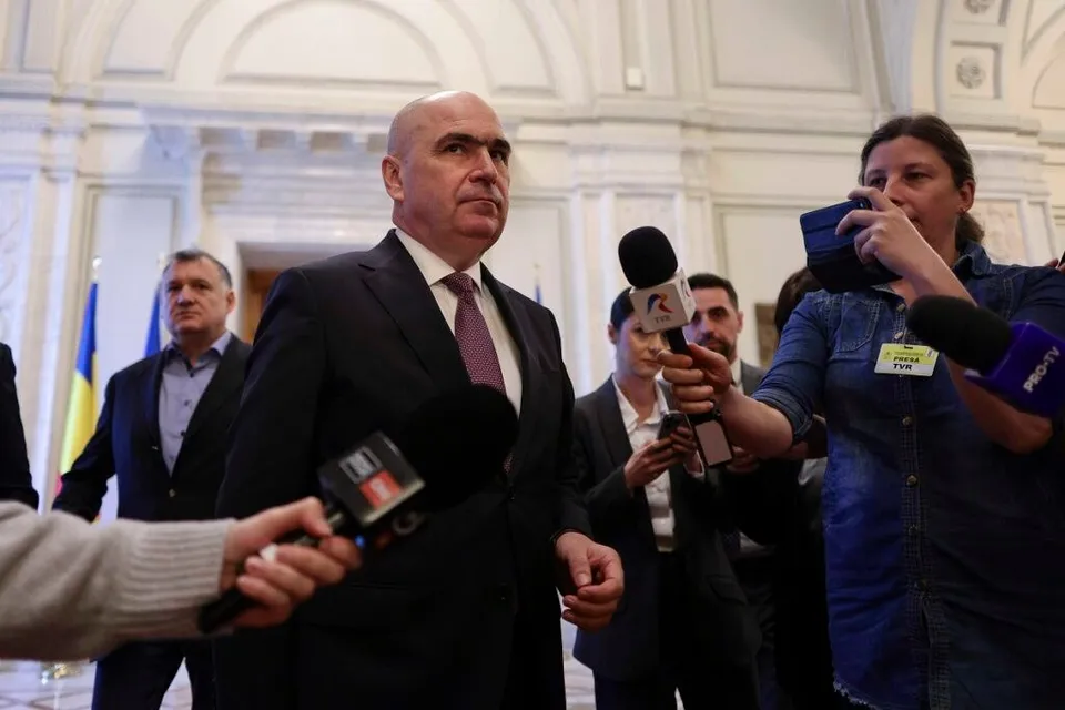 Un bărbat într-un costum închis și cravată purpurie este înconjurat de microfoane și persoane în interiorul unei clădiri cu arhitectură clasică.