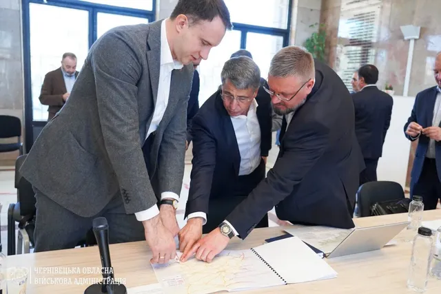 Trei bărbați în costume stau la o masă și privesc un document întins pe suprafața acesteia lângă un laptop și sticle de apă.