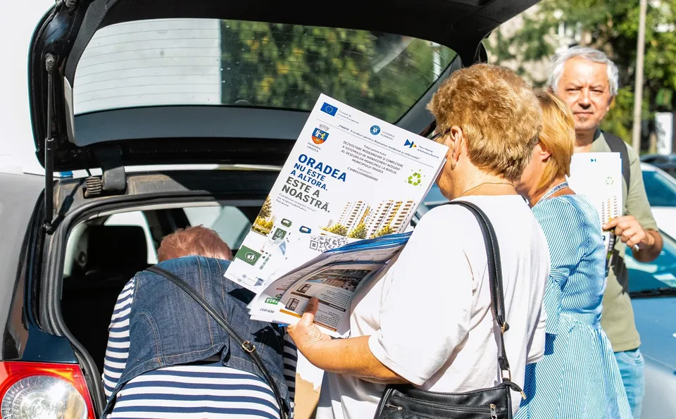 Trei persoane stau lângă portbagajul unui vehicul deschis, ținând în mâini diverse pliante și documente imprimate cu text și imagini colorate.