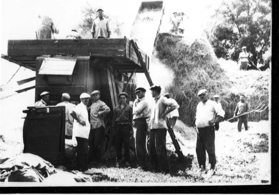 Un grup de bărbați stă lângă un utilaj agricol mare și o grămadă de fân sub un cer deschis.
