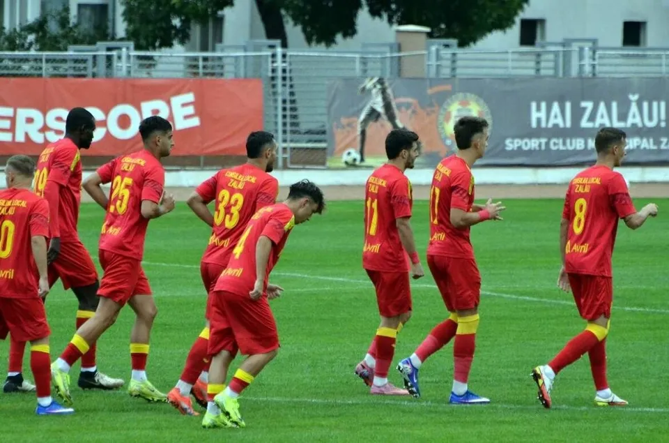 Câțiva jucători de fotbal îmbrăcați în echipamente roșii merg pe un teren de fotbal verde sub lumină artificială.