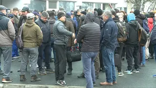Un grup de persoane stă adunat pe o stradă lângă o clădire, purtând haine de iarnă și rucsacuri.