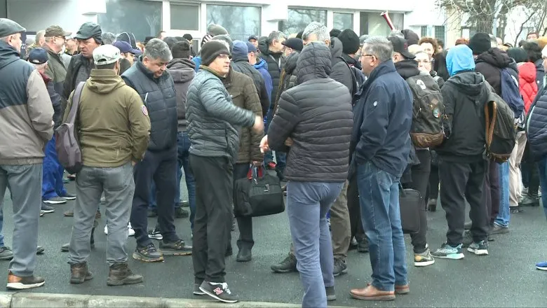 Un grup de persoane stă adunat pe o stradă lângă o clădire, purtând haine de iarnă și rucsacuri.