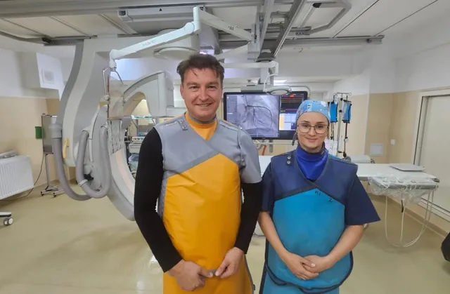 Un bărbat și o femeie poartă echipamente de protecție gri și colorate într-o încăpere medicală cu un aparat de radiologie în fundal.