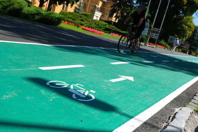 O persoană într-un tricou negru și pantaloni scurți circulă cu o bicicletă pe o pistă verde marcată cu simboluri și linii albe.