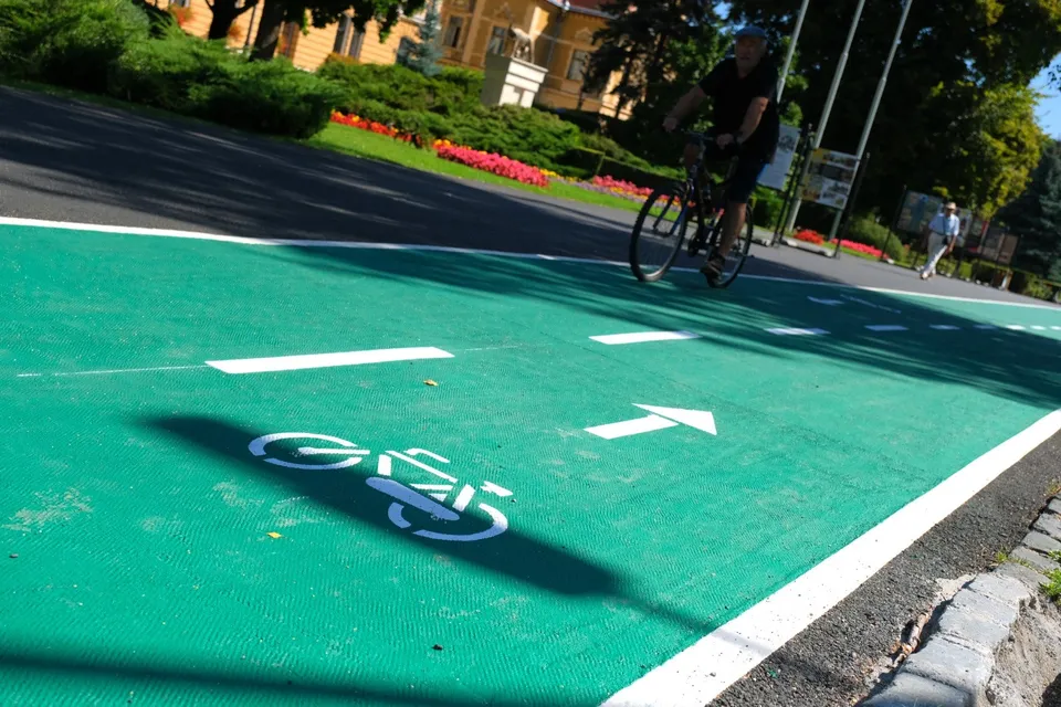 O persoană într-un tricou negru și pantaloni scurți circulă cu o bicicletă pe o pistă verde marcată cu simboluri și linii albe.