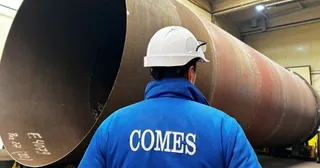 Un bărbat cu cască albă și haină albastră cu inscripția COMES privește un tub metalic de mari dimensiuni într-o fabrică.