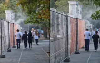Două persoane merg pe un drum asfaltat lângă un gard de plasă, în timp ce un nor de praf este vizibil în fundal.