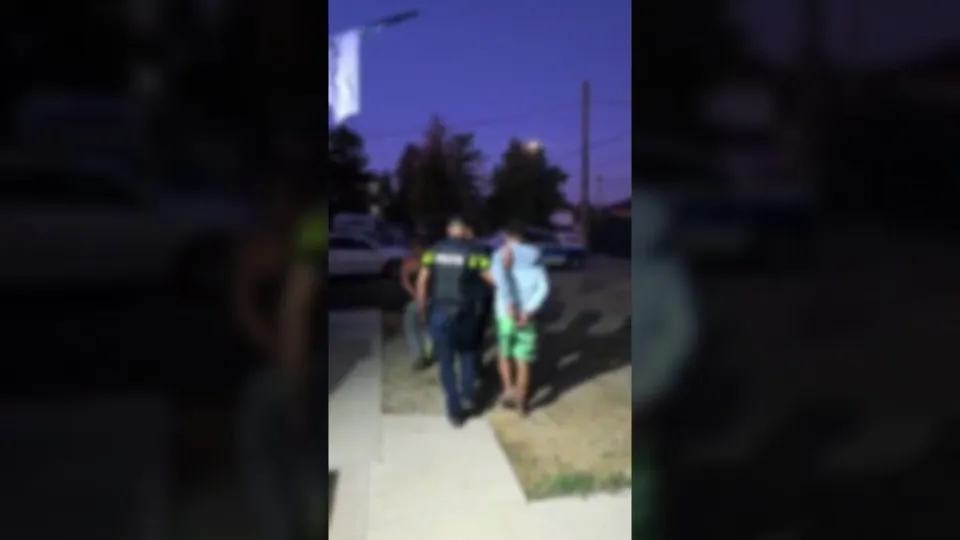 Un agent de poliție în uniformă întunecată conduce doi bărbați pe un trotuar în timpul serii.
