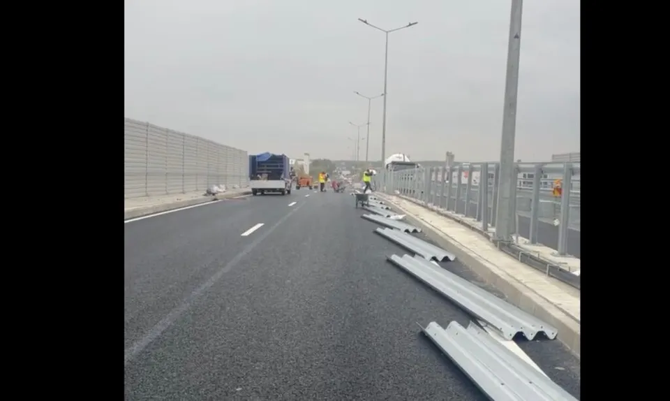 O porțiune de drum asfaltat cu bariere metalice de protecție pe marginea sa, un camion alb și stâlpi de iluminat sub un cer înnorat.