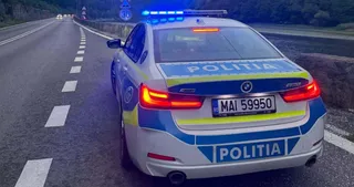 O mașină de poliție albă cu marcaje galben-albastre și lumini albastre aprinse este oprită pe marginea unei autostrăzi cu marcaje albe pe asfalt.
