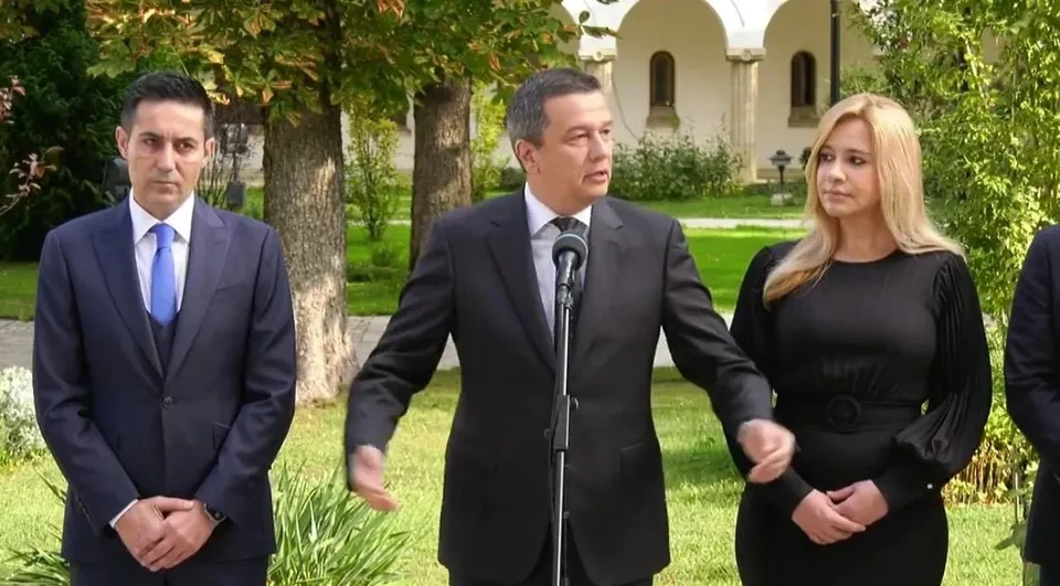 Un bărbat într-un costum negru vorbește la un microfon, fiind înconjurat de un bărbat în costum albastru și o femeie cu părul blond.
