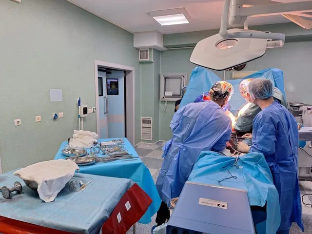 Echipe medicale în halate și mănuși sterile execută o procedură chirurgicală lângă un pacient acoperit în cadrul unei săli de operație.