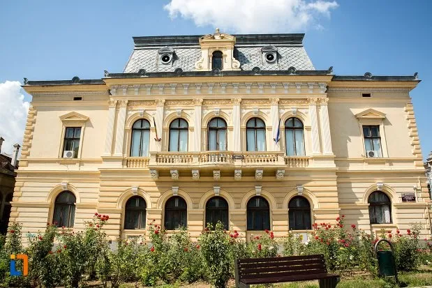 O clădire de două etaje cu fațadă galbenă, ferestre arcuite și un mansardă gri sub un cer cu nori.