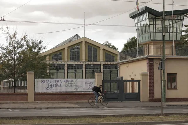 O persoană pe bicicletă trece pe lângă un clădire cu ferestre mari și un turn de observație sub un cer înnorat.
