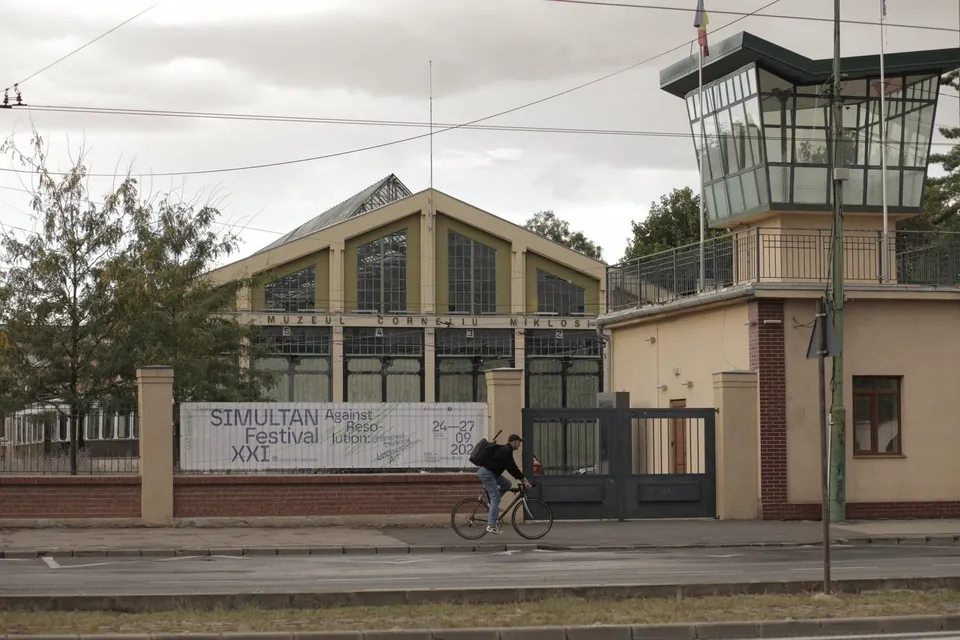 O persoană pe bicicletă trece pe lângă un clădire cu ferestre mari și un turn de observație sub un cer înnorat.