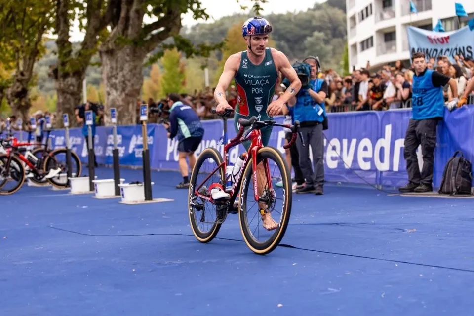 Un bărbat cu cască și echipament de triatlon merge pe o bicicletă roșie pe un covor albastru, în fața unui grup de spectatori.