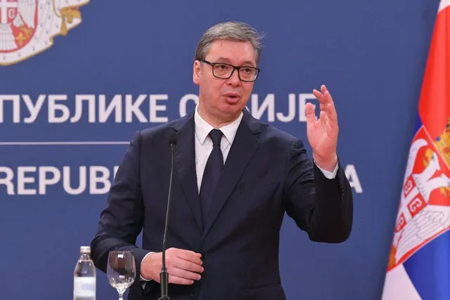 Un bărbat cu ochelari, purtând costum închis la culoare și cravată, stă în fața unui fundal albastru cu inscripții albe și un steag.