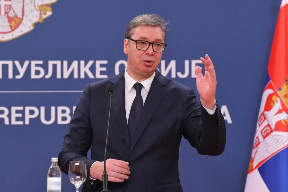 Un bărbat cu ochelari, purtând costum închis la culoare și cravată, stă în fața unui fundal albastru cu inscripții albe și un steag.