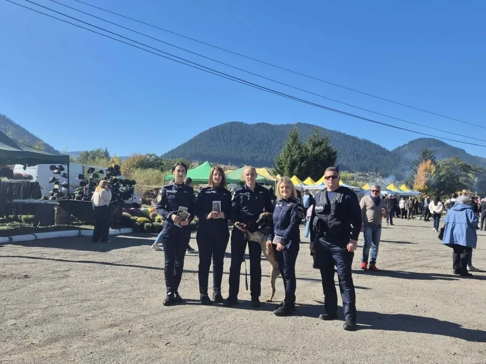 Cinci persoane în uniformă și haine civile stau în rând pe un teren deschis sub un cer senin, cu munți și copaci în fundal.